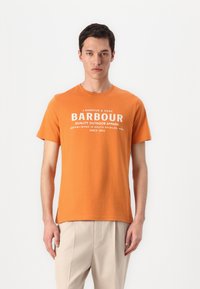Camiseta de algodão laranja com mangas curtas, decote redondo e texto impresso em branco destacando detalhes da marca e qualidade. Fendas laterais subtis.