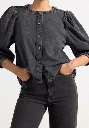 Person iført sort denimbluse med pufærmer og knapper foran samt matchende sorte højtaljede jeans, hænderne i lommerne.