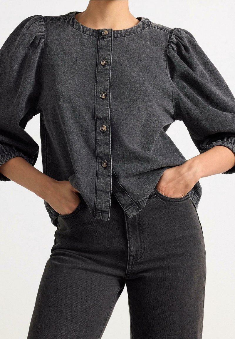 Personne portant un chemisier en denim noir à manches bouffantes avec des boutons sur le devant et un jean noir taille haute assorti, les mains dans les poches.