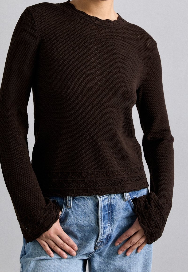 Brauner strukturierter Strickpullover mit langen Ärmeln, der einen gewellten Halsausschnitt und gemusterte Bündchen aufweist, kombiniert mit hellblauen Jeans.