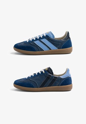 Deux baskets basses décontractées en daim bleu foncé et bleu clair avec des lacets blancs et des semelles en caoutchouc marron, vues de profil.