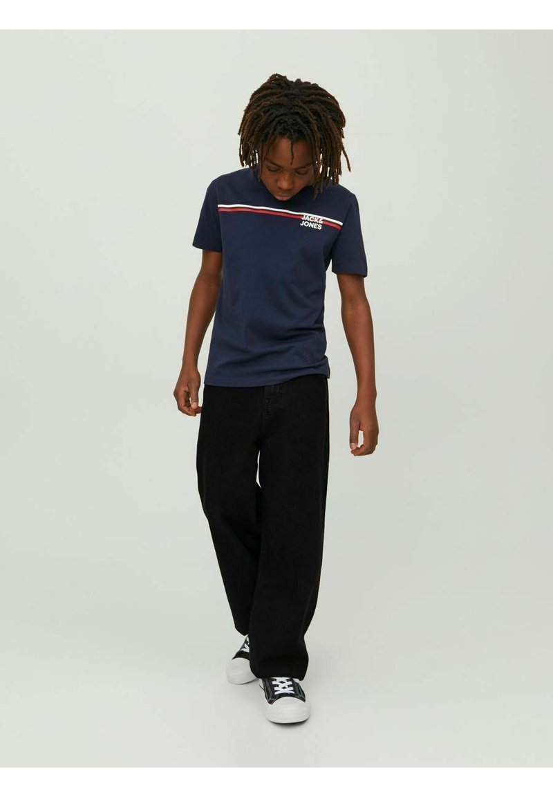 Marinblå bomullströja med röda och vita horisontella ränder och tryckt logotyp på bröstet, kombinerad med svarta vida byxor och sneakers.