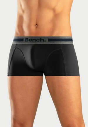 Männlicher Torso, der schwarze eng anliegende Boxershorts mit grauem Bund trägt, auf dem das Wort "Bench." steht, vor neutralem Hintergrund.