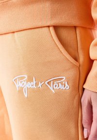 Orange bomulds joggers med ribbet talje, sidelomme og hvid broderet "Project x Paris" tekst på fronten.