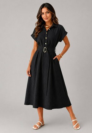 Femme portant une robe midi noire à manches courtes ceinturée avec des boutons dorés, des sandales blanches et des bijoux dorés, debout devant un fond uni.
