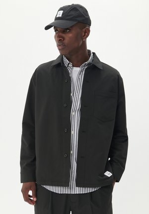 Black jacket with button closure, a chest pocket, and a smooth texture, layered over a striped shirt. Accented with a cap featuring a logo patch.