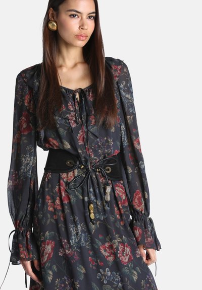 Femme portant une robe noire à fleurs à manches longues avec des poignets volantés et une large ceinture noire nouée à la taille avec des perles décoratives dorées et en pierre.