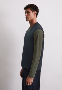 Dunkelblaue ärmellose Weste mit fünf Knöpfen, aus strukturiertem Stoff. Kombiniert mit einem langärmligen olivgrünen Hemd; das Modell ist im Profil dargestellt.