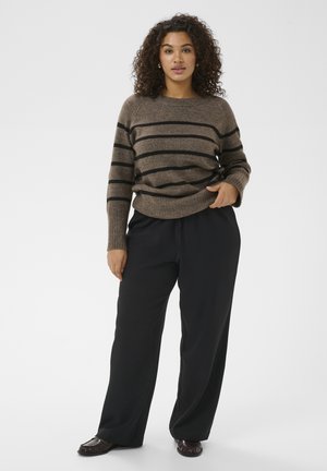 Kruhvliste juustega naine, kes kannab pruunide triipudega kampsunit, musta laia säärega pükse ja tumedaid mokasiine, seisab ühevärvilise tausta ees.