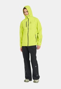 Veste imperméable jaune vif avec capuche, deux poches poitrine, deux poches latérales et fermeture éclair. Associée à un pantalon noir et des chaussures vertes.