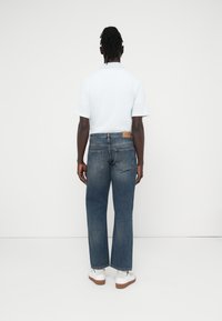 Μπλε τζιν παντελόνι με ίσια γραμμή, με μια ταμπέλα σε ταμπά χρώμα στο πίσω μέρος, συνδυασμένο με ανοιχτόχρωμο πουκάμισο με κοντά μανίκια και λευκά αθλητικά παπούτσια.