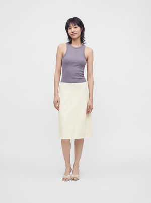 Joven mujer con camiseta sin mangas gris, falda crema hasta la rodilla y zapatos beige con punta abierta, de pie frente a un fondo blanco liso.