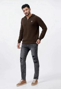 Maglione marrone a coste con colletto a mezza zip, abbinato a jeans grigi strappati e scarpe da ginnastica marrone chiaro. Logo prominente sul maglione.