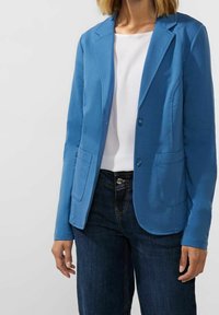 Blazer azul hecho de tela suave, con solapa con muesca, dos bolsillos delanteros y cierre de botones. Combinado con una camiseta blanca y jeans oscuros.