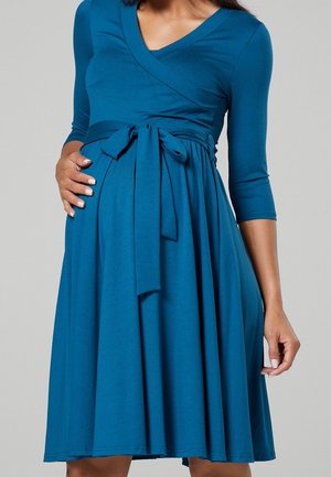 Teke kleurige wikkeljurk van zachte stof, met driekwart mouwen, een v-halslijn en een strikaccent aan de voorkant op de taille.