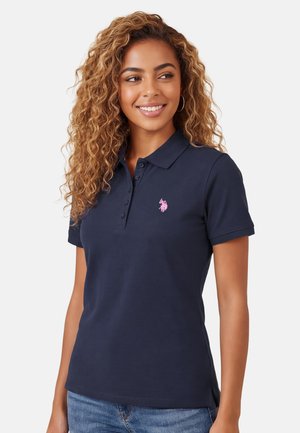 Woman with curly hair wearing navy blue polo shirt with pink embroidered logo, smiling and looking right, in front of a plain background.