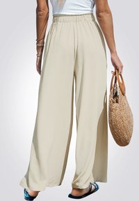 Pantalones de pierna ancha en tejido beige claro, con una cinturilla elástica, textura suave y un diseño fluido. Se sostiene un bolso de paja tejido.