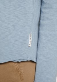 Světle modrá košile s dlouhým rukávem a strukturovanou tkaninou, s podšitým spodním lemem a malou etikettou s nápisem "Marc O'Polo Denim."