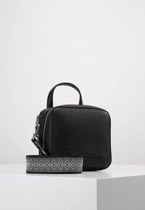 Sac à main noir en forme de carré avec une anse supérieure et une bandoulière amovible à motif noir et blanc, posé sur une surface blanche.