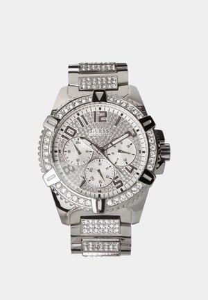 Sølv Guess-ur med krystalaccents, tre underskiver, numeriske timeangivelser og en metalrem indlejret med krystaller.