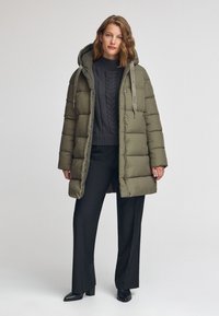 Olivgrön puffjacka med huva, parat med en mörk kabelstickad tröja och svarta byxor. Slät, quiltad textur, knappar fram.