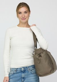 Bruine trekkoordtas van gladde stof, grote vorm met zijritsen, gedragen over de schouder. Gecombineerd met een witte top met lange mouwen en denim.