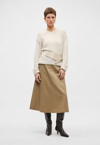 Krēmkrāsas džemperis ar ribotu aproci, jostas detaļa viduklī bēšā krāsā, brūna A formas svārki un melni, ceļgalu augstuma ādas zābaki ar papēdi.