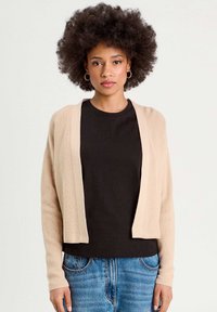 Cardigan en tricot beige avec des manches longues, design à devant ouvert, texture côtelée, porté sur un t-shirt noir, associé à un jean en denim bleu.