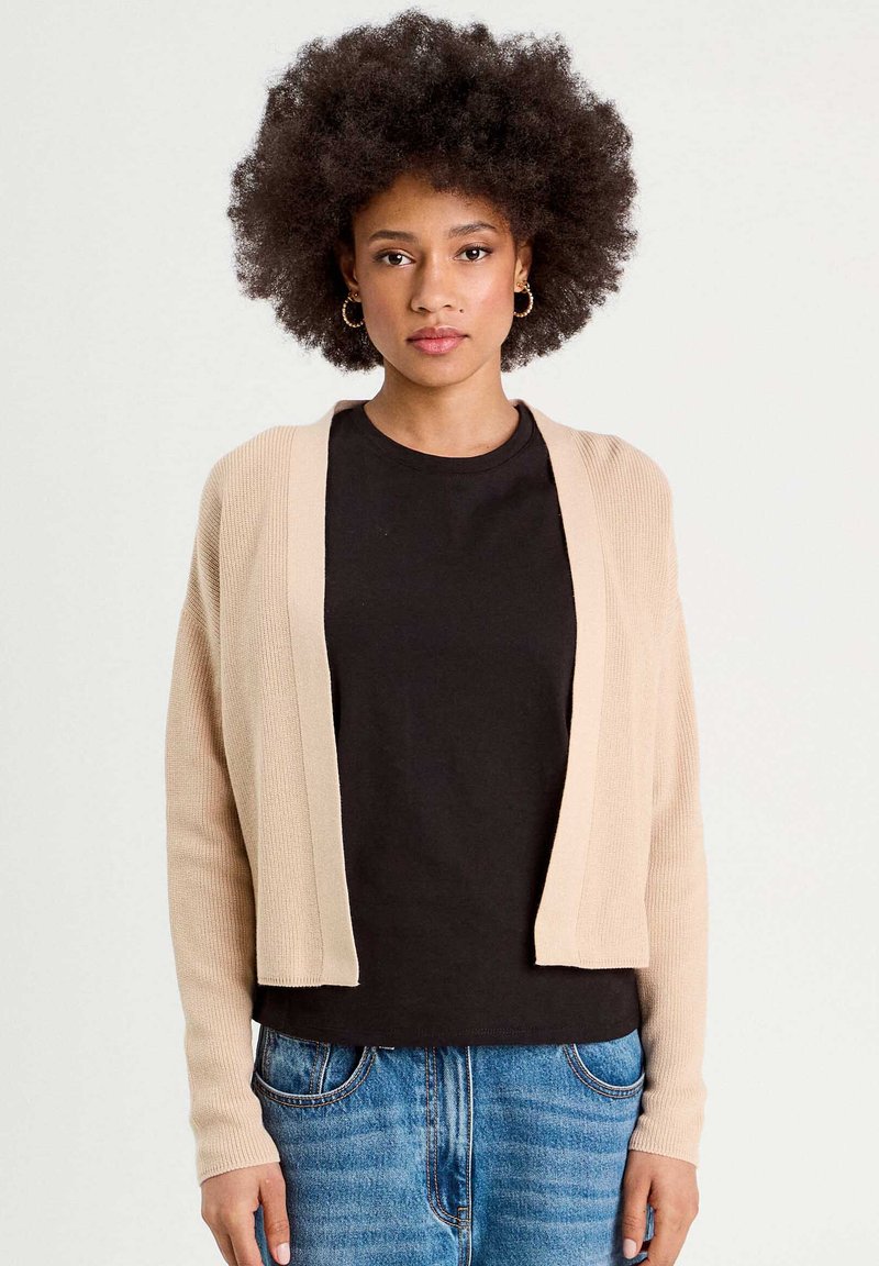 Cárdigan de punto beige con mangas largas, diseño de frente abierto, textura acanalada, usado sobre una camiseta negra, combinado con jeans de denim azul.