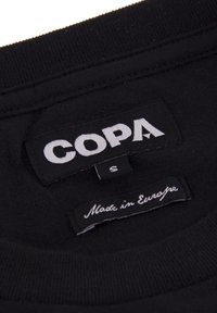 Zwarte stoffen label met "COPA" in witte tekst, maat "S" eronder, en "Made in Europe" in kleinere witte letters. Zachte structuur, standaard rechthoekige vorm.