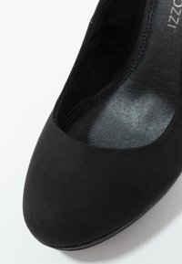 Chaussure à talon haut en daim noir avec un bout arrondi, une doublure intérieure lisse et peu de coutures apparentes. La semelle a une légère brillance.