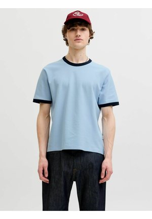 Młody mężczyzna w jasnoniebieskim T-shircie z ciemnymi lamówkami, ciemnych dżinsach i czerwonej czapce, stojący na jednolitym jasnym tle.