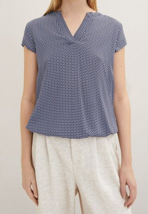 Kobieta ubrana w granatową bluzkę z krótkim rękawem z wzorem oraz jasnobeżowe spodnie z wysokim stanem, stojąca na neutralnym tle.