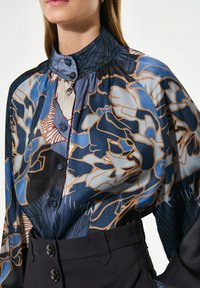 Sileda blouse, millel sinine lilleline mustriga, kõrge kraega ja nööbistamise ees. Koos kõrge vöökohaga mustade pükstega.