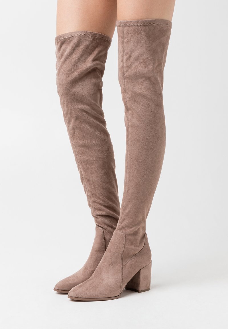 Stivali in suede alti fino alla coscia di colore taupe con tacchi a blocco indossati su gambe contro uno sfondo bianco semplice.