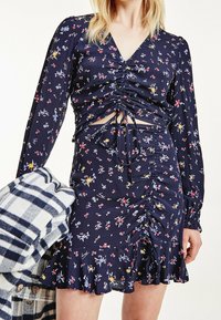 Abito mini floreale navy con maniche lunghe, dettagli arricciati, scollatura a V e orlo con volant. Abbinato a uno strato esterno a quadri grigio e bianco.