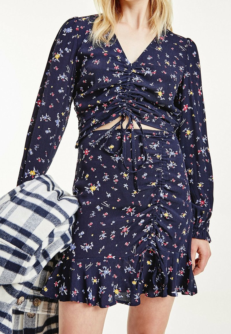 Abito mini floreale navy con maniche lunghe, dettagli arricciati, scollatura a V e orlo con volant. Abbinato a uno strato esterno a quadri grigio e bianco.