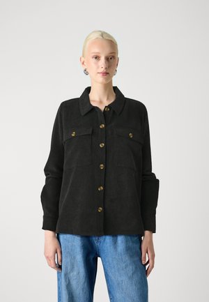 Black corduroy shirt with a collar, two front pockets, and brass buttons. Long sleeves with rolled cuffs, paired with light blue denim pants.