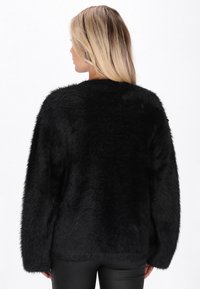 Donna con lunghi capelli biondi che indossa un maglione nero morbido a maniche lunghe e pantaloni neri, vista da dietro su uno sfondo bianco.