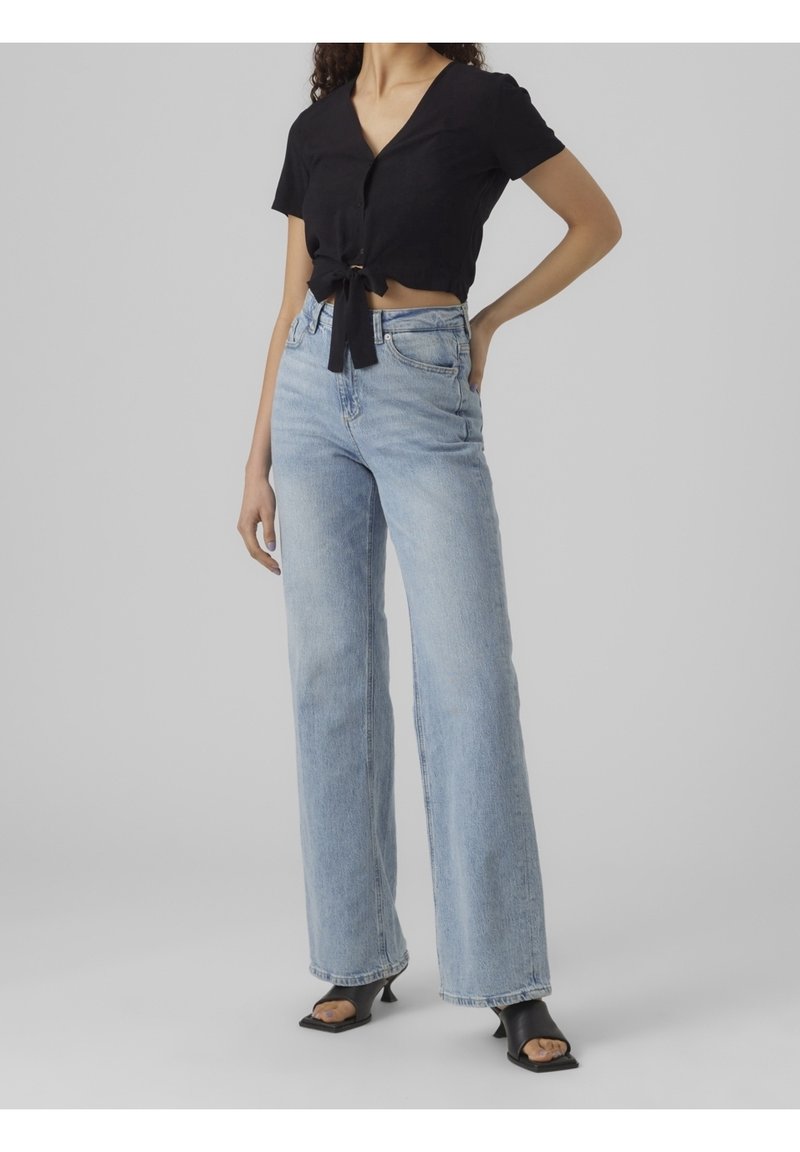 Femme portant un jean bleu clair taille haute à jambes larges, un top noir à manches courtes noué devant, et des sandales à talons noires sur fond gris.
