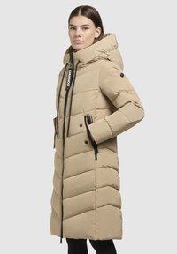 Manteau en duvet beige avec un col montant, couture en chevrons verticales, zippers visibles, poches latérales et capuche avec cordon de serrage.