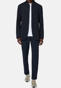 Mörkblå blazer med mandarin-krage och tygknappar, ihop med matchande byxor och en vit skjorta. Skorna är ljusgrå med svarta accenter.