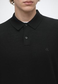 Homme portant un polo en maille noire avec deux boutons et un petit logo "cK" sur le côté gauche de la poitrine.