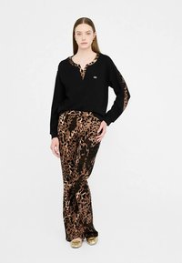 Zwarte top met lange mouwen en V-hals met een goudkleurig logo, gecombineerd met wijde broek met luipaardprint in bruin en zwart. Gouden schoenen maken de outfit compleet.