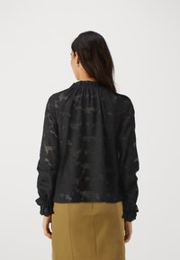 Blusa nera trasparente con motivo floreale, scollatura arricciata e maniche lunghe con polsini. Indossata con una gonna marrone chiaro, che mostra una vestibilità rilassata.