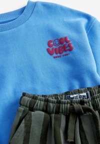 Sweatshirt bleu avec le texte "COOL VIBES" et un motif de smiley. Pantalons de jogging rayés olive avec taille élastique et détail à nouer.