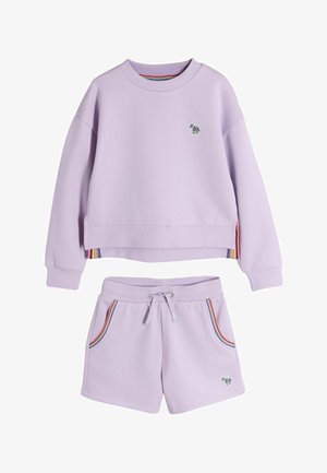 Mladé dievča s dlhými vlnitými vlasmi, ktoré má na sebe svetlofialovú mikinu a zodpovedajúce šortky so malými zebrovými nášivkami, usmieva sa do kamery.