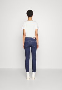 Jeans ajustados de mezclilla azul oscuro, con bolsillos traseros cosidos, talle medio, y una camiseta blanca lisa con mangas cortas arriba.