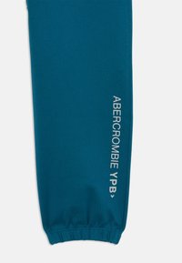 Teal jogger pants made of soft fabric with an elastic cuff. Features white print detailing on the left side reading "ABERCROMBIE YPB."