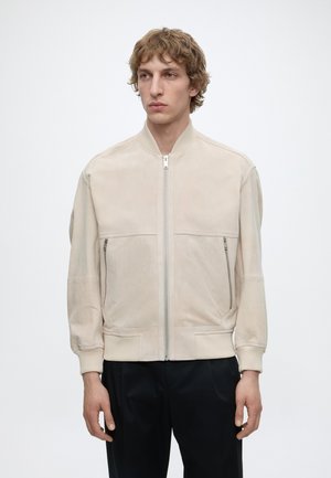 Jeune homme aux cheveux bouclés portant une veste bomber beige zippée avec des poches zippées et un pantalon noir, debout devant un fond uni.